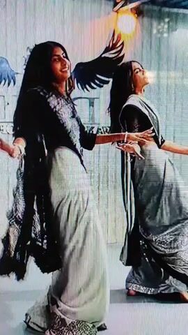 Two women in saris are dancing. The woman on the left is wearing a black blouse and a light-colored sari with a black and silver border. The woman on the right is wearing a black blouse and a gray sari with a sparkly border. They are both in motion, with their arms extended. The background appears to be a wall with some decorative elements, including what looks like a bird with outstretched wings and a light fixture that resembles a flame. The lighting is somewhat dramatic, with bright spots and shadows.