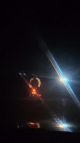A very dark video shows what appears to be a person on a motorcycle from behind, with their taillight visible. The camera is moving, and there are bright lights in the distance, possibly from other vehicles, creating glare and lens flare effects. The overall scene suggests movement at night on a road.