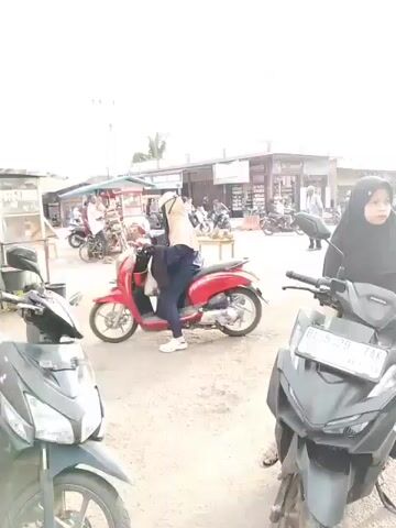 The video appears to be shot from a moving motorcycle, looking forward and slightly to the right. The scene is an outdoor street with several shops and market stalls lining both sides. A variety of motorcycles are parked and in motion. In the foreground, a dark-colored scooter is visible. Further ahead, a red scooter is ridden by a person wearing a hijab. A woman in a black hijab is also on a scooter, partially obscuring the view of the red scooter. Beyond them, a white car is driving down the street. The sky is overcast, and there are utility poles and wires visible overhead. The overall impression is a busy, everyday street scene in a town or city.