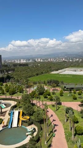 The video showcases an expansive view from an elevated position, likely a balcony or high-rise window, capturing a cityscape framed by a partly cloudy blue sky. In the foreground, there's a water park area with multiple pools, colorful water slides, and landscaped grounds featuring walking paths, grass, and palm trees. Beyond the park, a lush green space extends toward the city buildings, which are arrayed against a backdrop of distant hills or mountains. The camera pans to reveal a large apartment complex. The pools in the water park are empty, and the overall scene appears serene and devoid of people.