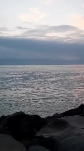 The video shows a body of water with small waves under a cloudy sky. The foreground consists of dark rocks. The scene is mostly still, with only the movement of the waves. The overall lighting suggests either dawn or dusk.