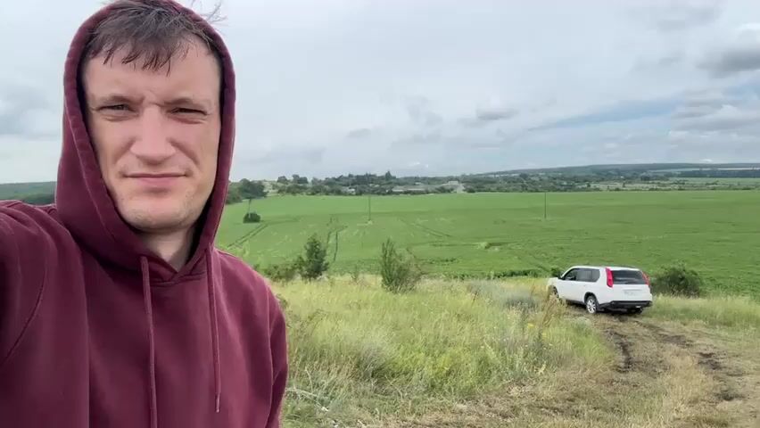 A man in a maroon hoodie is filming himself with a camera, taking a selfie. He is standing in a grassy area on a slight incline. Behind him, a white SUV is parked on a dirt track, facing away from the camera. The landscape behind the SUV is characterized by rolling green fields, scattered trees, and a cloudy sky. The man appears to be looking directly at the camera with a neutral expression.