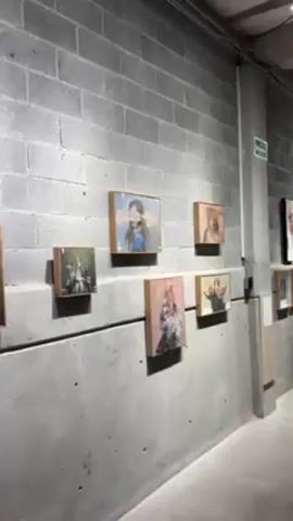 The video shows a collection of framed paintings displayed on a light gray concrete block wall. The paintings are hung in a linear fashion, with a slight vertical offset between the rows. The paintings are primarily portraits with blurred or impressionistic features. A piece of white paper with handwritten notes and a lime green sign with white text are affixed to the wall at the side. A small table with papers or pamphlets is near the far end of the wall. The room is well-lit and appears to be an art gallery or studio space.
