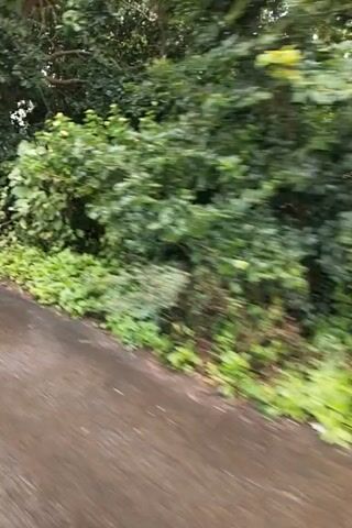A blurry, vertical video shows a person's perspective walking along a wet, paved road. Lush green foliage lines the left side of the road, with some areas appearing overgrown. A corrugated metal structure is visible among the greenery, and further down the road, there are piles of debris, including plastic bags and a blue barrel. The right side of the road appears to be a dirt path or shoulder, also wet. Beyond the foliage, a field with visible rows of crops can be seen, and the sky is overcast. The footage is shaky, suggesting movement, and the overall environment appears rural and somewhat unkempt.