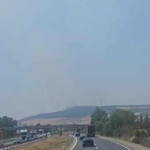 The video shows a hazy sky over a rural landscape. In the distance, a hill is visible with several wind turbines on its crest. Smoke plumes are rising from the hillside, indicating a fire. In the foreground, a highway overpass is present with traffic moving on it, including trucks and cars. Trees and fields are also visible in the foreground and middle ground. The overall impression is one of a hot, dry environment possibly affected by a wildfire.