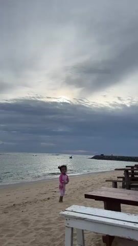 A young girl in a pink swimsuit stands on a sandy beach facing left, with her back to the ocean. She is looking towards a body of water with waves. In the background, a breakwater extends into the ocean. Further back, several people are seated at picnic tables. The sky is cloudy with some sunlight breaking through. Wooden picnic tables are in the foreground.