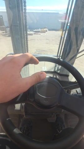 The video shows a first-person view from inside the cabin of a construction vehicle. A black steering wheel is prominently in the foreground. The person operating the vehicle is visible by their hand on the steering wheel and their feet in sneakers at the bottom of the frame. Through the windshield, a cluttered construction site is visible, with scattered materials and equipment on a dirt surface. A large, light-colored building stands in the background under a blue sky.