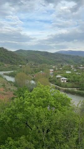 The video shows a landscape view of a small village nestled in a valley. A river flows through the valley, and lush green hills rise on either side. The sky is overcast with a mix of clouds and some blue sky. The village consists of scattered houses, mostly one or two stories, with red or gray roofs. The surrounding area is heavily wooded with various trees and vegetation. There's a road or path running parallel to the river on one side. The camera seems to be positioned on higher ground, looking down on the village. The general impression is one of tranquility and natural beauty.