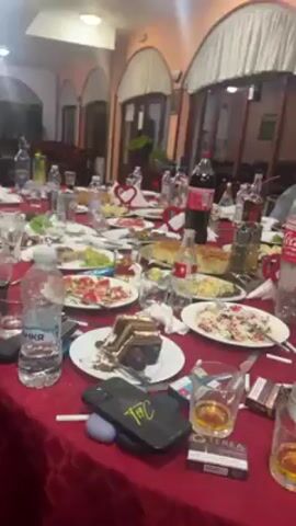 The video shows a post-meal gathering at a restaurant or banquet hall. A long table is covered with a red tablecloth and laden with used plates, leftover food, open bottles of liquor and soda, cigarette packs, and personal items like a smartphone and wireless earbuds. Some people are seated around the table while one man is playing bagpipes. The room is decorated with arched window-like structures covered in white curtains.  The lighting is bright.
