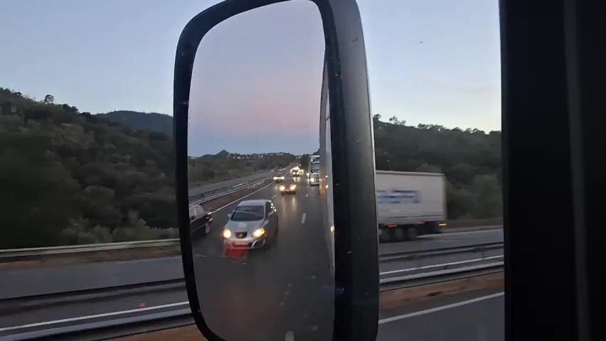 The video shows a rear-view mirror reflection of a highway at dusk. The sky is a light purple and blue, with the moon visible in the upper left corner. The highway has multiple lanes with cars and trucks driving on it. Headlights are visible from the approaching vehicles. In the foreground of the mirror, a silver car with its headlights on is being driven by a person wearing a red shirt. To the left of the mirror, a white truck is seen driving on the highway. The background of the mirror shows a hilly landscape covered with trees. The right side of the video shows the side of the truck cab and a blurry view of the highway and trees outside the truck.
