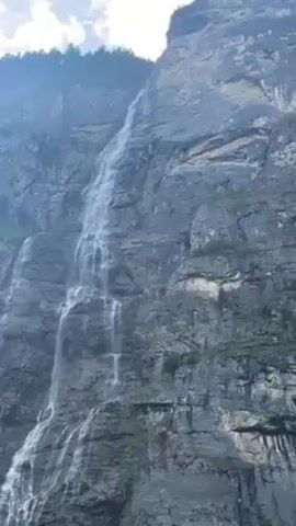 The video shows a tall, rocky mountain with a waterfall cascading down its side. The waterfall has multiple tiers as it descends, and there are patches of green vegetation growing on the mountain's surface. The sky above is partly cloudy.