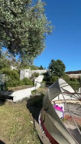 The camera pans up from ground level, revealing a deteriorating structure with metal supports and remnants of white fabric. The structure is on a concrete platform covered in faded square tiles. Beyond the platform, a body of water is visible, with a dense forest on the opposite shore. The sky is clear and blue. The overall scene appears to be an abandoned recreational area with a view of the water. There are remnants of graffiti on the structure. The scene is overgrown with vegetation.
