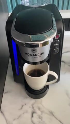 A video captures a Monarchy Coffee Roaster machine brewing coffee into a white mug placed on a marble countertop. The coffee drips steadily into the mug, filling it with a dark liquid. The machine is black with silver accents and features a blue light on its side. The background shows a light blue tiled wall.