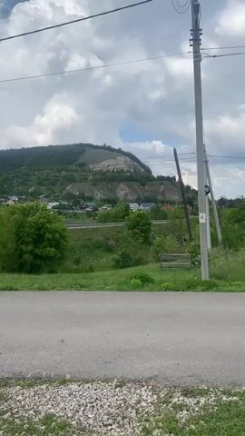 A still shot of a rural landscape with a road in the foreground, a grassy slope, and a village nestled at the base of a lush green mountain range under a cloudy sky. A telephone pole stands to the right of the frame.