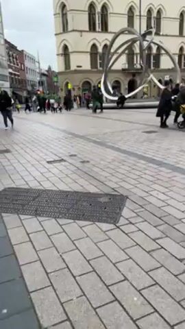 The video shows a city square, paved with light gray bricks and featuring a large, abstract, silver-colored sculpture in the center. People are walking around the square, some with strollers. The buildings surrounding the square are multi-storied and made of brick, some with white accents. The sky is overcast. Some individuals are dressed in green, possibly for a St. Patrick's Day celebration. There is a manhole cover visible in the foreground. The camera moves slightly as the videographer walks.