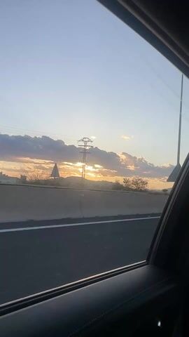 The video, shot from inside a moving vehicle, shows a sunset viewed through the car window. The sun is partially obscured by dark, puffy clouds, with golden rays streaming through the gaps. In the distance, mountains can be seen silhouetted against the bright sky. A highway barrier runs along the bottom of the frame, and power lines are visible overhead. The car's interior is dark, providing a contrasting frame for the vibrant scene outside.
