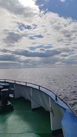 The video captures a boat moving across a large body of water during the daytime. Initially, the shot shows the front of the boat with its green deck and blue railing, looking out over the water towards the horizon under a partly cloudy sky. The sun is visible through the clouds, reflecting on the water. The camera then pans to the stern, revealing the frothy white wake created by the moving boat. The dark blue water is rippled, and the horizon line shows distant land. The overall scene conveys a sense of peaceful transit on a vast waterway. No text is visible.