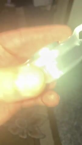 The video shows a close-up of a metallic object being held in a hand. The object appears to be a component with intricate detailing, including numbers and possibly markings like "28" and "1xc tic". It has a central opening and several indentations and raised sections around it. A small tab or lever protrudes from the side. The hand holding the object is visible from the fingertips to the palm. The lighting suggests an indoor environment, possibly dimly lit, with a blurred background that hints at furniture or a patterned surface. The object is rotated to show different angles, highlighting its construction and markings.