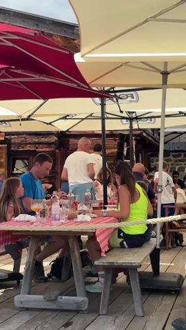 The video shows an outdoor seating area at what appears to be a restaurant or cafe. Several tables with red and white checkered tablecloths are set up on a wooden deck. People are seated at these tables, some eating and drinking, others conversing. In the foreground, a group of people are sitting at a picnic-style table. A man in a blue t-shirt and a young girl are on the left side of the table, while a woman in a neon yellow top is on the right. A man in a white t-shirt is standing in the background, interacting with someone at another table. There are umbrellas providing shade over some of the tables. In the background, a rustic-looking building with wooden walls and stone accents is visible, suggesting a mountain or alpine setting. The overall atmosphere is casual and relaxed, with people enjoying a meal outdoors.