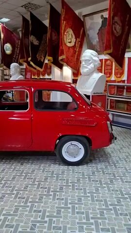 This is a video of a museum exhibit that appears to be dedicated to Soviet history. A small, red car is parked in the foreground, facing right. Behind the car, there are two motorcycles, one blue and yellow, and another black. The walls are adorned with portraits of historical figures, including a large portrait of Vladimir Lenin. There are also several red flags and banners, some with inscriptions. Busts of historical figures are also visible. The floor is tiled in a geometric pattern. The lighting is bright and even, suggesting an indoor setting. The video pans slowly to the right, offering a view of the exhibit from different angles. There are no people visible in the video.