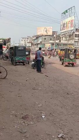 The video depicts a street scene in what appears to be a South Asian town. The ground is dirt and uneven, with visible trash scattered around.  Several auto-rickshaws, some covered with green canvas tops, are parked on the side of the road. A few buses and other vehicles can also be seen, suggesting a busy transportation hub. People are present, some walking, some riding motorcycles, and one person on a bicycle. Overhead, power lines crisscross the sky. Several billboards featuring women's faces and product advertisements are visible on the buildings in the background. The overall impression is of a bustling, slightly chaotic street scene.