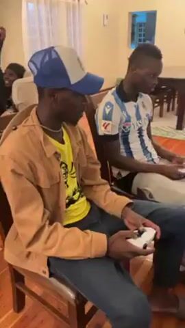 Two young Black men are seated and playing a video game using white controllers. The first man is wearing a yellow t-shirt, a brown jacket, a blue hat, and blue jeans. The second man is wearing a blue and white striped jersey, and light-colored jeans. They are sitting in a living room with wooden floors, and a couch with a small screen in a carrying case on it, which shows the game they are playing. There is a table and chairs visible in the background. The lighting is warm and interior.