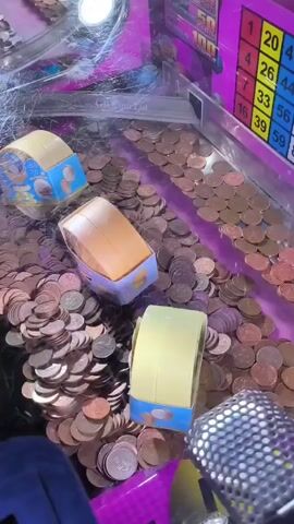 The video shows a close-up view of a coin-operated arcade game, likely a push-penny or coin pusher machine. The machine is filled with a large quantity of copper-colored coins, mostly pennies, forming a dense layer across the playing surface. Three small, cylindrical objects, possibly prizes or targets, are scattered amongst the coins. These objects have a light brown or orange top and a light blue base with images of coins on them. In the foreground, a dark purple, textured area of the machine is visible, with a slot opening for coin insertion. To the right, a portion of a pink game board with a grid of numbers and colors is visible, suggesting a bingo or lottery-style element to the game. The lighting is somewhat dim and purple-hued, typical of arcade environments. The camera angle is from above, looking down into the machine. There are no people visible in the frame. The coins appear to be in disarray, with some piled higher than others.