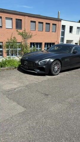 A dark gray Mercedes-Benz sports car is parked in front of a brick building with modern white and gray sections. The car is viewed from the side, with its front end slightly angled towards the camera. The car has silver alloy wheels with red brake calipers visible behind them. The building has several windows and a large exhaust pipe extending from the roof. The sky is blue and clear, indicating a sunny day. The ground is asphalt, with some cracks and debris visible. The video appears to be shot from a moving vehicle or a handheld camera, as there is slight movement throughout.