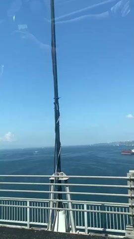The video shows a view from a bridge overlooking a body of water. The sky is blue with some white clouds. There are white railings in the foreground, and large concrete and metal structures of the bridge are visible. In the distance, there are boats and land. The camera appears to be moving, as the perspective shifts slightly throughout the short clip.