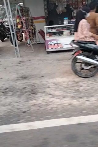 The video shows an outdoor scene. In the foreground, a white and red structure with an electronic display is visible. To the left, a train track and some trees can be seen. The ground is a mixture of dirt and concrete. Further into the scene, a motorcycle is parked. In the background, there are buildings and stacks of lumber. The video appears to be shot from a moving vehicle, likely a car. The orientation of the video is sideways.