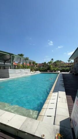 A vertical, eye-level video shows a bright, sunny day with a clear blue sky. The main focus is a swimming pool filled with turquoise water. The pool has a white tiled edge and a wooden deck on the right side. On the left side of the pool, there is a covered area with white furniture and some stacked white materials. In the distance, there are buildings and lush green trees and palm trees. A person is visible swimming in the pool, with their head above water. The camera is positioned at the edge of the pool, looking across its length.