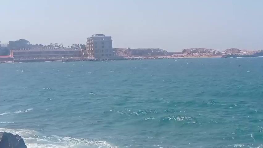 The video opens with a wide shot of a body of water, likely the sea, with waves lapping against rocks in the foreground. In the distance, a complex of buildings is visible along the coastline, with trees dotting the landscape behind them. The sky is clear and hazy, suggesting a warm, sunny day. As the video progresses, the camera pans to the left, revealing a concrete walkway or path along the water's edge. A person wearing a white top and pink shorts is walking along this path, facing away from the camera. Further along the path, two people are seated on a bench or low wall, their backs to the camera. The waves continue to crash against the rocky shore, creating a dynamic and lively coastal scene. The buildings in the background appear to be a mix of modern and older structures, some of which might be hotels or residences. The overall impression is of a pleasant day at the seaside.