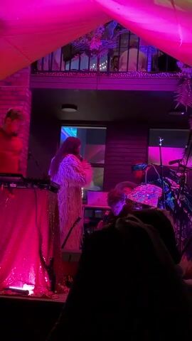 A live music performance is captured from a low angle. A woman with long dark hair, wearing a fuzzy, white, floor-length coat, stands center stage holding a microphone and singing. To her left, a man in a red hoodie is operating a DJ setup with a table covered in a sparkly pink cloth. To the woman's right, another person, wearing a hat and dark clothing, is seated and also holding a microphone. Behind them, a drummer is visible playing a drum set, with cymbals reflecting the pink and purple stage lights. The stage is dimly lit with vibrant pink and purple hues from overhead lighting and accent lights. In the background, a balcony area with several people is visible, also illuminated by the colored lights. The overall environment appears to be an indoor venue, possibly a bar or club, with brick walls and a ceiling draped with fabric and plants.