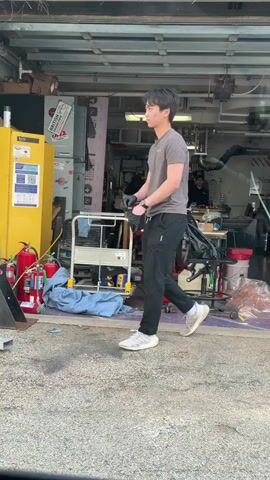 The video shows two young Asian men in a garage workshop. The first man is wearing a gray t-shirt, black pants, and white sneakers; he enters from the left, puts on black gloves, and walks to the right. The second man has glasses, a similar gray t-shirt, gray sweatpants, and white sneakers with yellow accents. He is wearing the gloves as he walks in from the left, stopping in the center, and he talks while gesturing with his hands. Three people are working at a long table in the background of the garage. The garage is cluttered with tools, a red gas can, and a red fire extinguisher. A large white sign with black lettering is visible at the back right of the garage. The floor is gray concrete.
