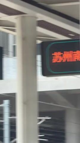 The video shows a train station platform. There are signs indicating platform numbers. Sign 3 is visible, and a digital sign with Chinese characters is also present. People are seen on the platform, some with luggage, and a train is visible in the background. The platform is paved with tiles and there are overhead structures and electrical lines for the train. The overall environment appears to be a modern train station.