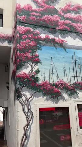 A medium shot shows a mural of a flowering tree with pink blossoms on the exterior wall of a building. To the left, a woman in a pink shirt is taking a photo with her phone. She is standing in a walkway with brick paving and people walking in the distance. Above the walkway entrance, there is a rectangular mural depicting sailboats in a harbor. A tall, multi-tiered planter filled with greenery and flowers is located to the left of the woman, partially obscuring the mural. The camera pans slightly to the right, revealing more of the flowering tree mural and a window with a Firefox logo. The environment appears to be a pedestrian area, possibly a shopping arcade or promenade.