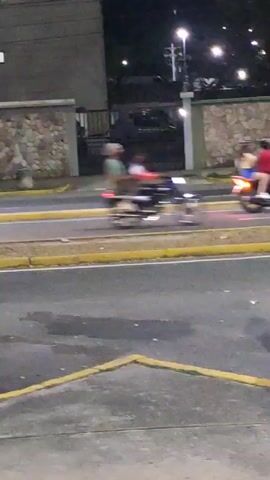 The video shows a street scene at dusk. A blue car is parked on the left side of the frame behind a yellow curb. In the background, there is a low stone wall and a building. Several motorcycles speed by on the street, moving right to left. The video is a bit blurry.
