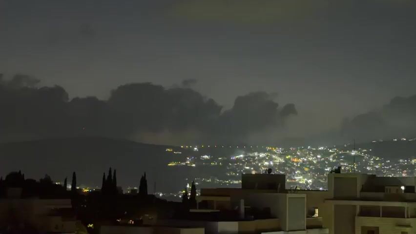 This is a nighttime aerial shot of a city. The sky is dark with some clouds, and a city lights glow in the distance. In the foreground, there are several buildings with lights on. There are also some trees and other greenery. The video appears to be a time-lapse, as the clouds and city lights seem to be moving slightly. There are no people or animals visible in the video.