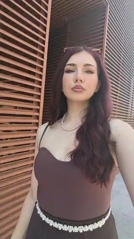 A young woman with long, dark auburn hair, styled in loose waves, is seen from the chest up in a video selfie. She is wearing a brown, strappy tank top with a sweetheart neckline and a white beaded belt. She also wears a white pearl necklace and sunglasses resting on top of her head. Her makeup includes winged eyeliner, eyeshadow, and lip gloss. The video shifts from an indoor setting with a brown slatted wall to an outdoor garden with green grass and trees under a clear sky. The woman looks directly at the camera with a neutral expression throughout the video, which is shot from a slightly low angle.