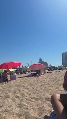 The video shows a crowded beach on a sunny day. The sand is a light brown color and there are many beach umbrellas spread out, in various colors including pink and green. People are sitting and lying down on the sand, some under the umbrellas for shade. In the background, there is a clear blue sky, some buildings, and what appears to be industrial chimneys or towers. There is a person's leg visible in the foreground, suggesting the video is taken from a seated perspective on the beach.
