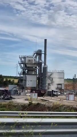 The video shows a construction site with an asphalt mixing plant. There are several trucks on site, some of which appear to be loading or unloading materials. The plant has tall silos and structures associated with industrial operations. The environment is somewhat barren with exposed earth and sparse vegetation. The sky is cloudy. The camera appears to be moving, possibly from a vehicle.