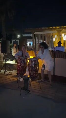 Two women are playing conga drums at night outdoors. The woman on the left is wearing a light-colored top and the woman on the right is wearing a floral-patterned robe over shorts. There is a smaller drum in front of the conga drums. In the background, there is a raised wooden structure with lights and people sitting at tables. There is also a palm tree and a speaker on a stand to the left of the frame.
