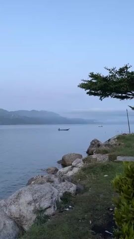 The video displays a serene natural landscape, focusing on a body of water with mountains in the distance. The foreground features a rocky shoreline with grass and a tree branch extending from the right side of the frame. The water is calm, reflecting the pale blue sky and misty mountains. A small, narrow boat with a single occupant is visible in the middle of the lake, slowly moving from left to right. Another similar boat is further in the distance on the right. The overall atmosphere is tranquil, suggesting either dawn or dusk due to the soft, diffused light and muted colors.