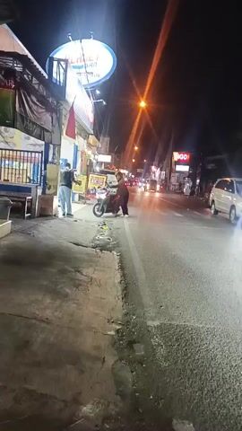 The video shows a street scene at night. Several motorcycles are visible, some with riders. There are buildings lining the street with illuminated signs and storefronts. A few cars are also present, with headlights and taillights visible. The overall impression is of a busy street in an urban area at night.