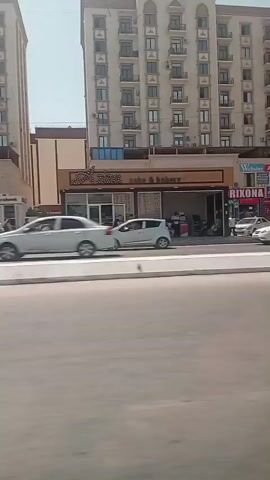 The video shows a street scene in Termez, Uzbekistan on a sunny day. A tall, modern apartment building with many balconies dominates the background. Below the apartment building are several storefronts. A white car drives on the road in the foreground.

Visible text includes:
- "CAKE & BAKERY"
- "WEBSTER ACADEMY"
- "DORIXONA" (pharmacy)
- "APPLE TRADE"
- "MADINA FARM"
- "ZARGAR"
- "AN OR MEN'S"
- "WHITE HOUSE BOUTIQUE"

People are visible in front of some of the shops, and a bus with colorful advertisements is also seen driving on the street. The overall impression is of a bustling urban environment.
