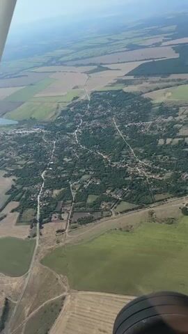 An aerial drone shot captures a sprawling rural village nestled amidst agricultural fields. The village is densely packed with houses, many of which appear to have red-tiled roofs, interspersed with numerous trees, creating a lush green canopy. Several roads and pathways wind through the settlement, connecting individual homes and leading out towards the surrounding countryside. The agricultural landscape is a patchwork of different crops, with fields of varying colors and textures, suggesting a diverse range of agricultural activity. In the distance, more rolling hills and fields stretch towards the horizon, bathed in the soft light of a hazy sky. Part of an aircraft wing and tire are visible in the foreground, indicating that the footage was likely captured from a low-flying plane.