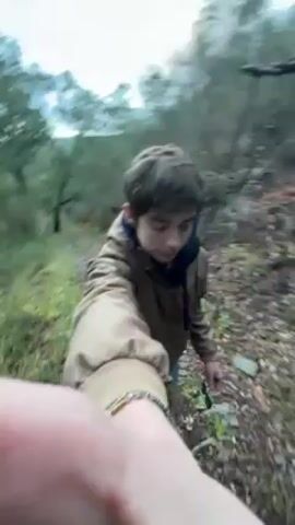 A young person, possibly a teenager, is recording a video while walking on a narrow trail in a wooded area. The trail is lined with trees and vegetation. The person is wearing a brown jacket, a dark scarf, and jeans. The camera is held in front of the person, with their hand partially visible in the foreground. The sky is overcast. The person says "Let's go" as they continue walking.