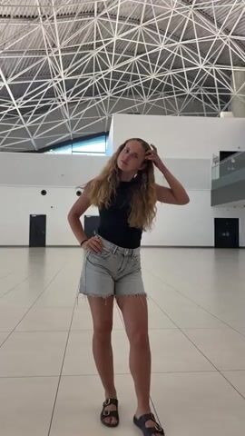 A young woman with long blonde hair stands in a large, well-lit room with a high, angular ceiling. She is wearing a black tank top, light denim shorts, and black sandals. The floor is tiled in a light color with dark grout lines. There are doorways visible in the background, as well as a multi-level structure with railings. The woman poses for the camera, touching her hair and shifting her weight. She turns her back to the camera and then faces it again, her body in motion. The video appears to be shot from a low angle.