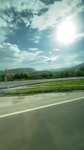 The video shows a view from inside a moving vehicle, traveling on a highway. The sun shines brightly in a partly cloudy sky. Green grass and trees line the side of the highway. There is a blurred view of a car moving in the same direction on the adjacent lane. The highway has a guardrail.
