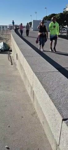 A group of people walk along a paved path next to a concrete seawall. The path is curved and slopes upwards from left to right. The sun is shining, casting long shadows. The person furthest to the right is wearing a bright yellow jacket and dark shorts and appears to be jogging. Next to them, a person wearing a dark tank top and patterned leggings walks while talking on a phone. Further ahead, two people in casual clothing walk side-by-side. One person is wearing a pink shirt and a hat, and the other is wearing a grey shirt and a hat. In the distance, another person in blue is standing on the edge of the path. To the left of the path, there is a beach with some beach chairs and a tent. Trees and buildings are visible in the background.
